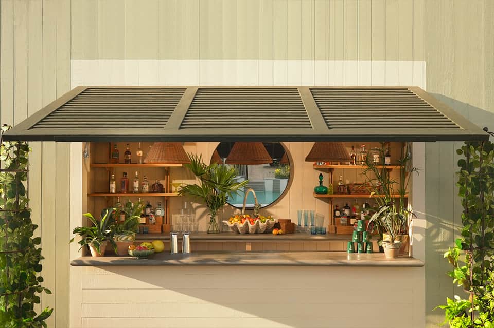 Outdoor bar counter with wooden shelving, bottles, plants, round mirror, pendant lights, and retractable awning