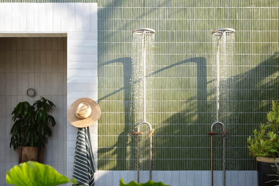Two outdoor shower fixtures with green corrugated wall backdrop and tropical plantings