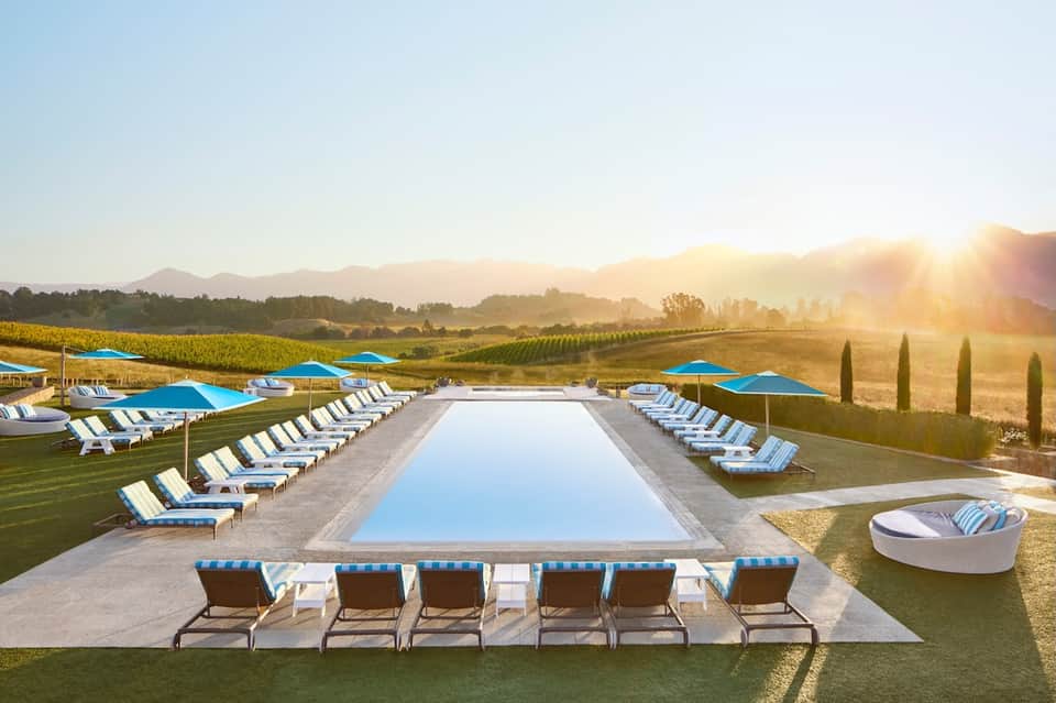 Resort lap pool with blue umbrellas and loungers overlooking vineyards and mountains at sunrise