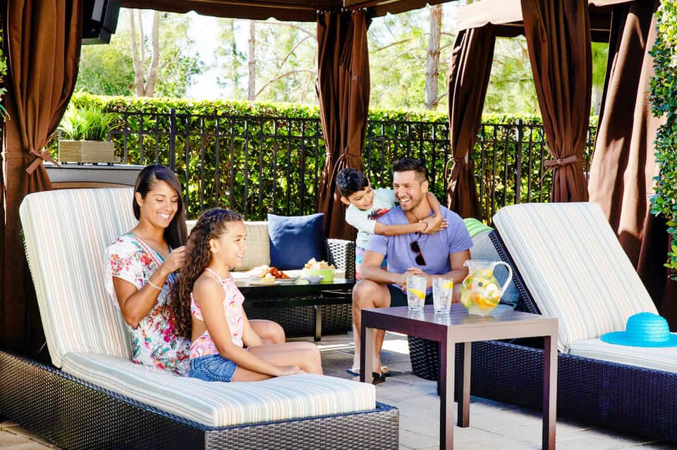 Family of four relaxing on lounge chairs under brown cabana with striped cushions and curtains
