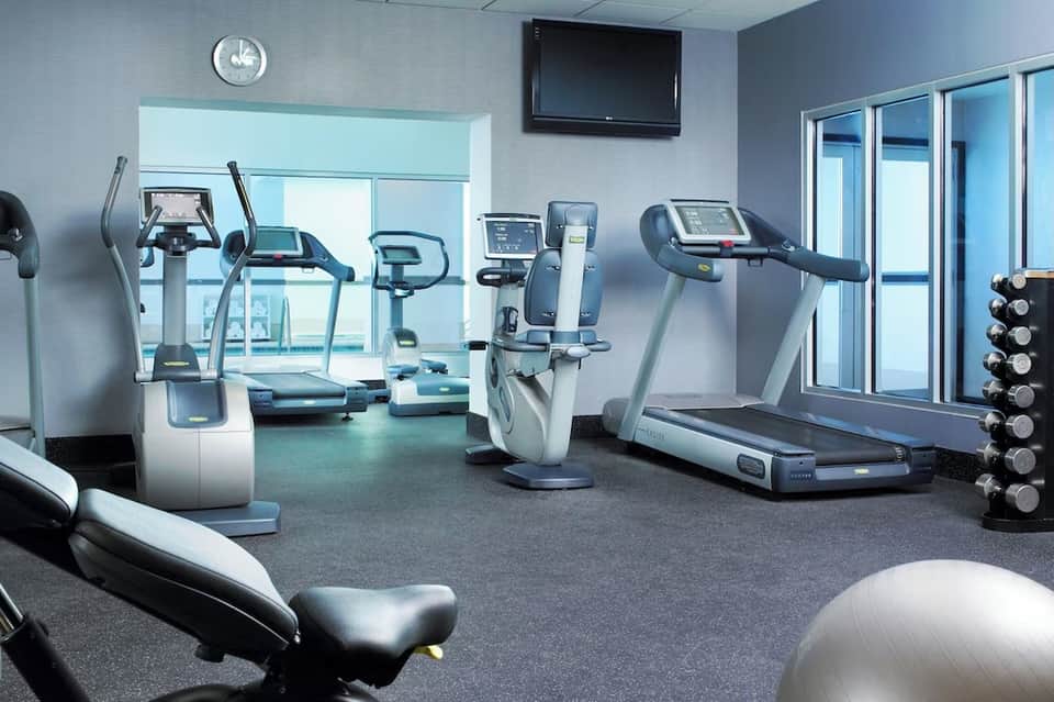 Modern fitness center with treadmills, stationary bikes, dumbbells, and large windows with aqua tones