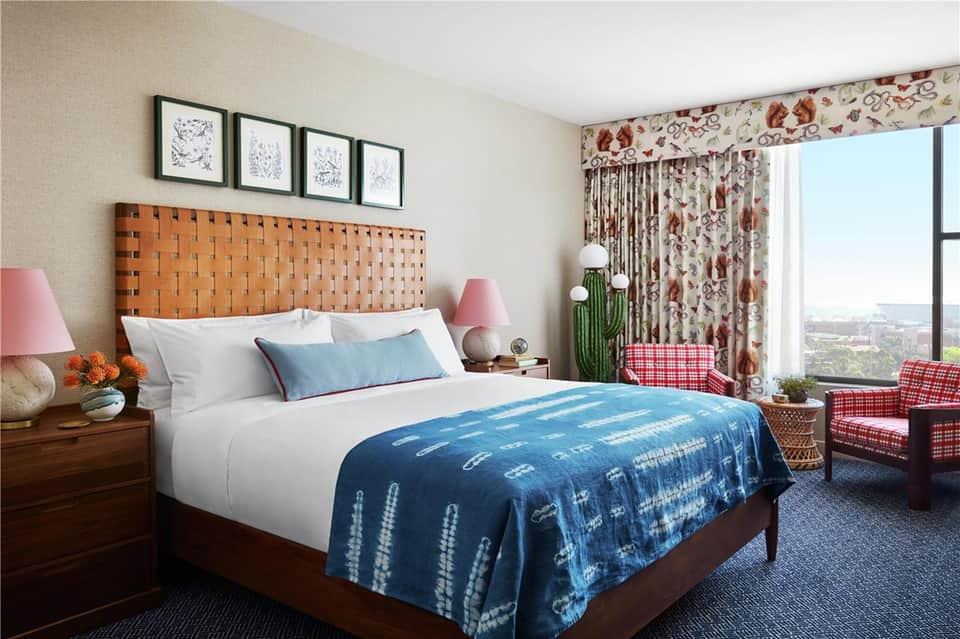 Guest bedroom with orange woven headboard, white bedding, blue throw blanket, floral curtains, and city view