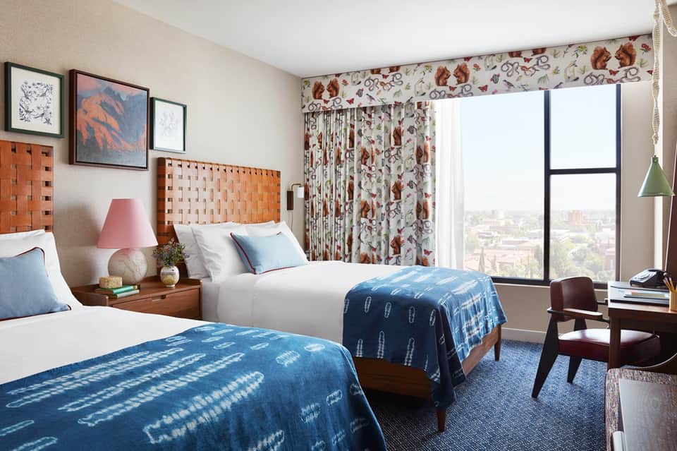 Upscale hotel room with two beds, woven headboards, floral curtains, and city views
