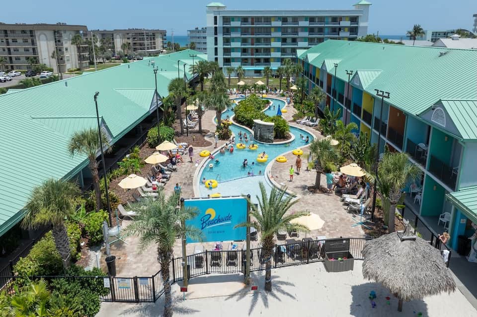 Aerial view of beachfront resort with curved pool, colorful buildings, palm trees, and ocean
