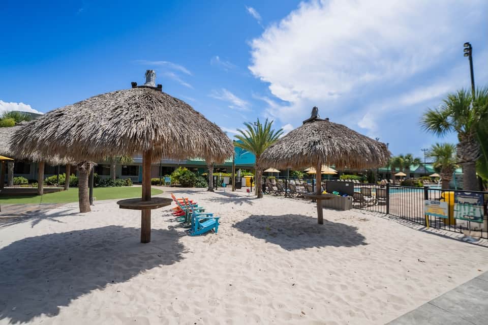 Beach area with thatched-roof palapas, fire pit, lounge chairs, and palm trees by ocean