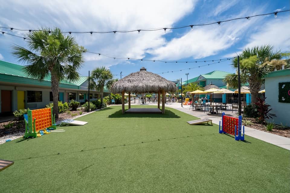 Colorful resort grounds with green lawn, tiki hut, palm trees, and activity areas