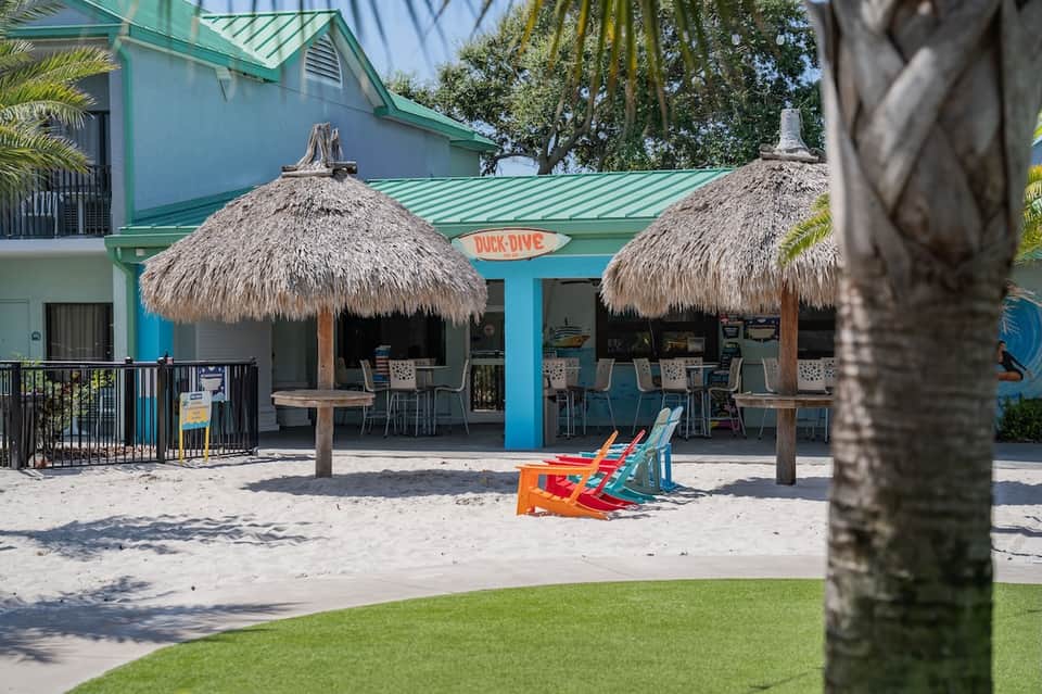 Beachfront bar with thatched roof umbrellas, turquoise building, and colorful beach chairs on sand