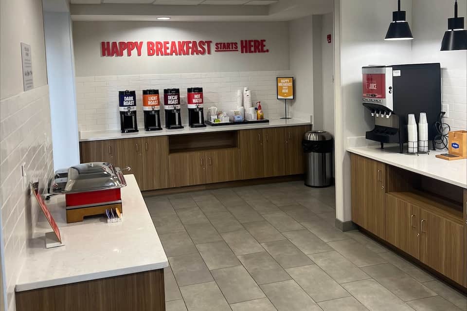 Hotel breakfast area with coffee dispensers, signage reading 'Happy Breakfast Starts Here'