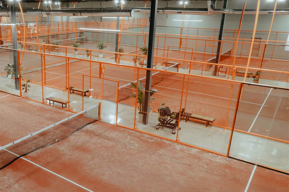 Indoor tennis facility with multiple red clay courts separated by orange safety barriers under lights