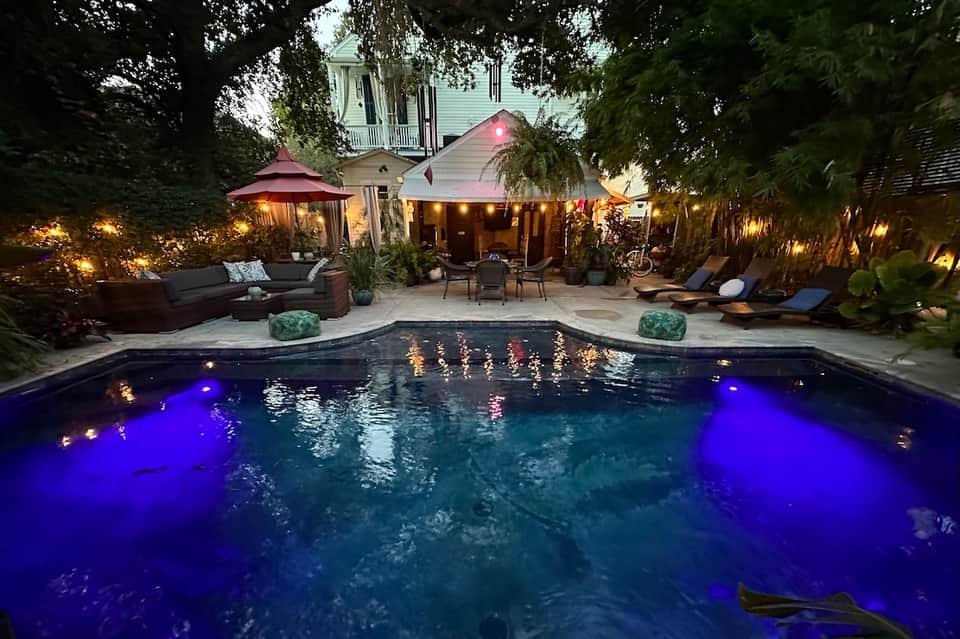 Backyard pool illuminated with blue lights at dusk, surrounded by lounge seating and trees