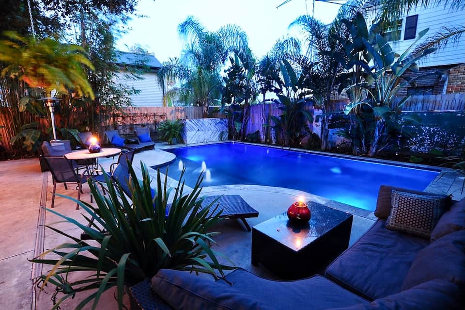 Illuminated rectangular pool at night surrounded by tropical plants, lounge seating, and a fire pit