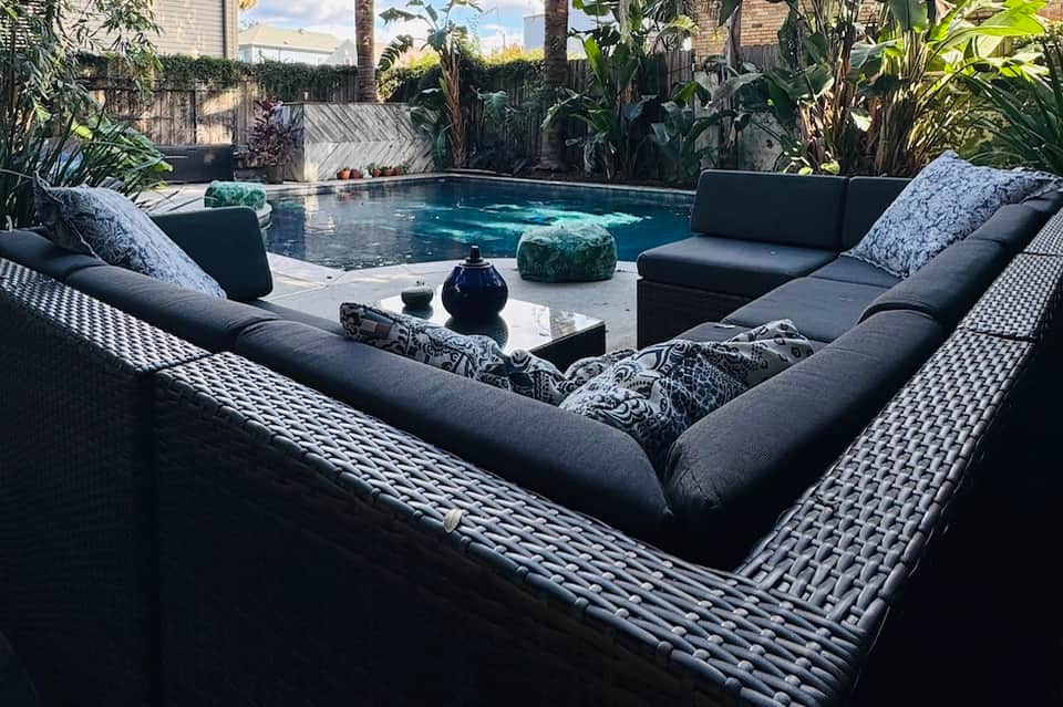 Modern backyard pool surrounded by tropical plants, black lounge furniture, and stone decking
