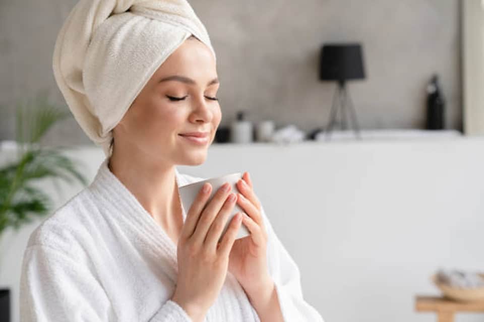 Woman in white spa robe and towel wrap holding warm beverage in modern bathroom setting