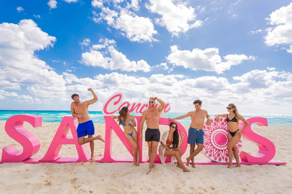 Group of people posing with pink Cancun sign letters on sandy beach with turquoise ocean