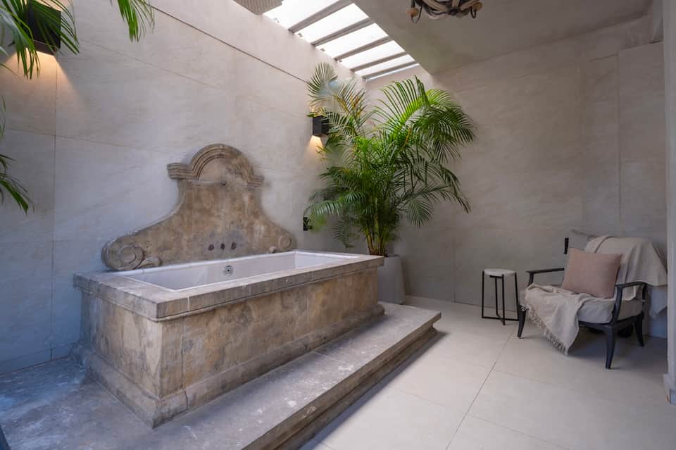 Spa bathroom with ornate stone soaking tub, skylight, potted palm, and lounge chair