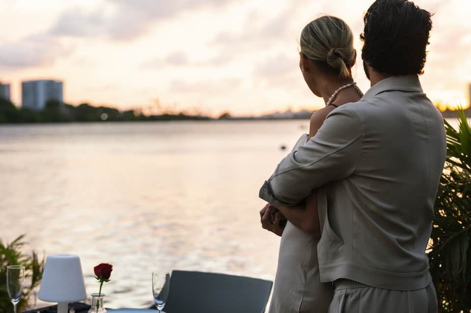 Couple embracing on waterfront balcony at sunset with city skyline reflected in water
