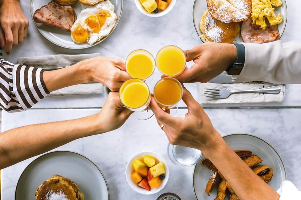 Overhead view of brunch table with four hands holding glasses of orange juice, pastries, eggs, and fresh fruit