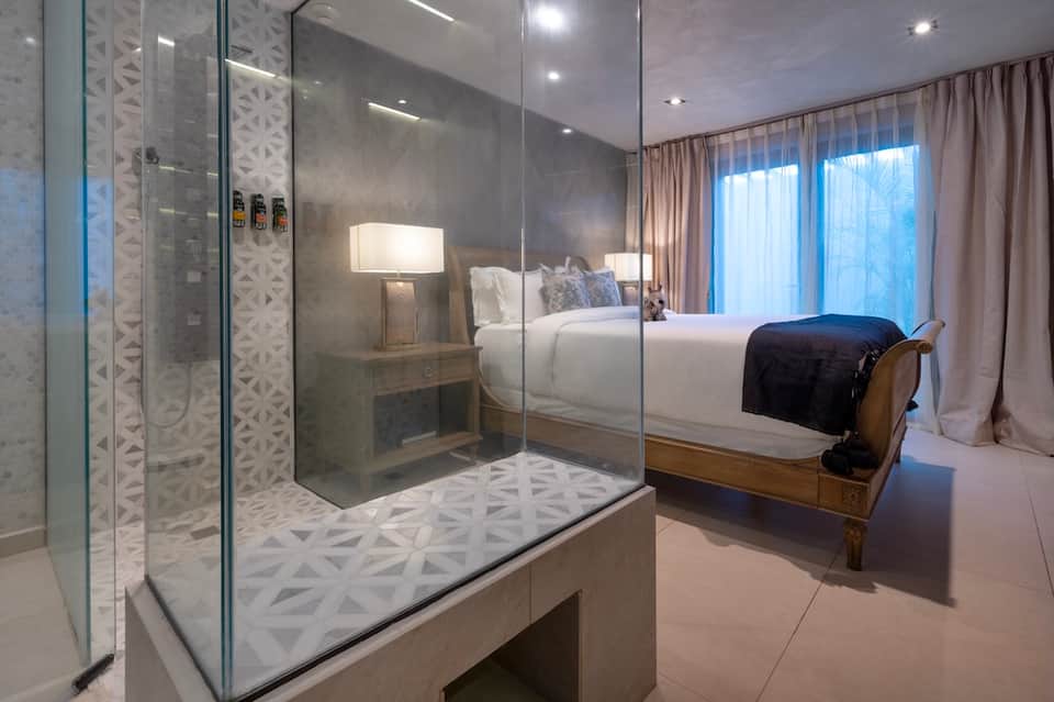 Modern hotel bedroom with glass shower enclosure, white bedding, and floor-to-ceiling windows