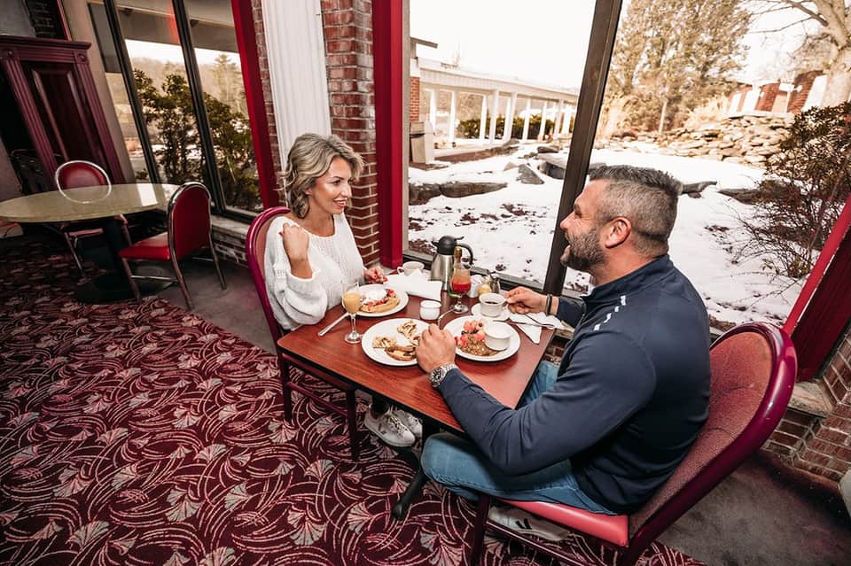 Couple dining at hotel restaurant with large windows, burgundy accents, and outdoor winter garden view
