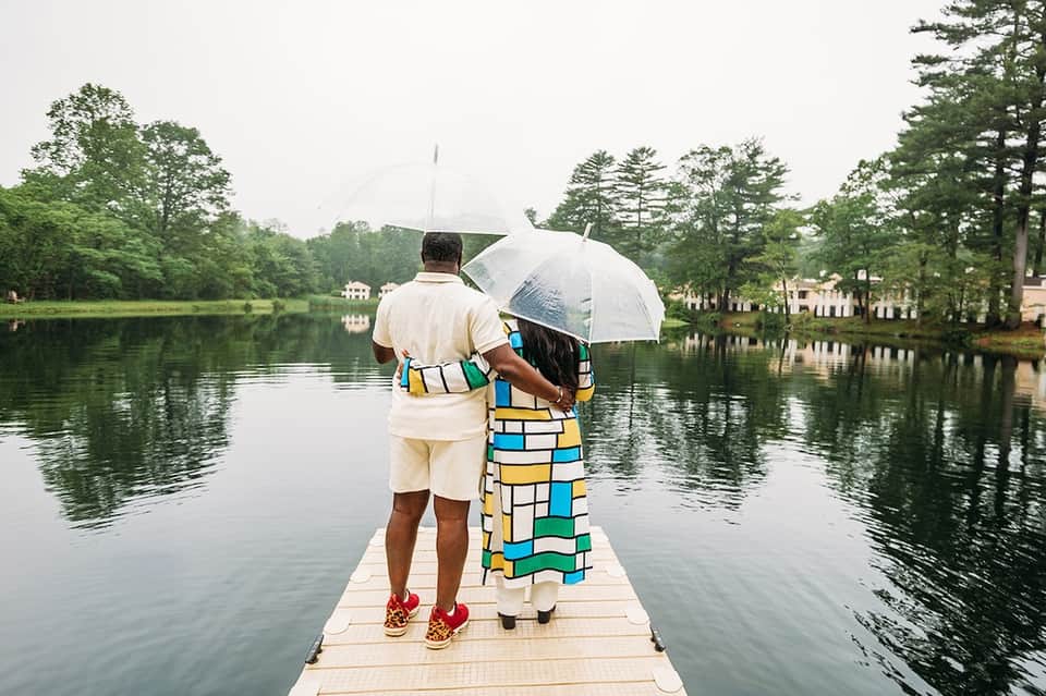 Couple with umbrella standing on dock overlooking calm lake surrounded by lush green trees and homes