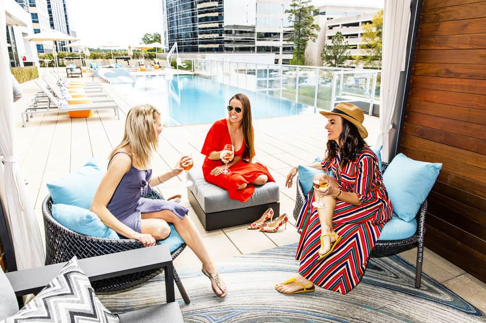 Three women enjoying cocktails on a rooftop lounge with a resort pool and city skyline in background
