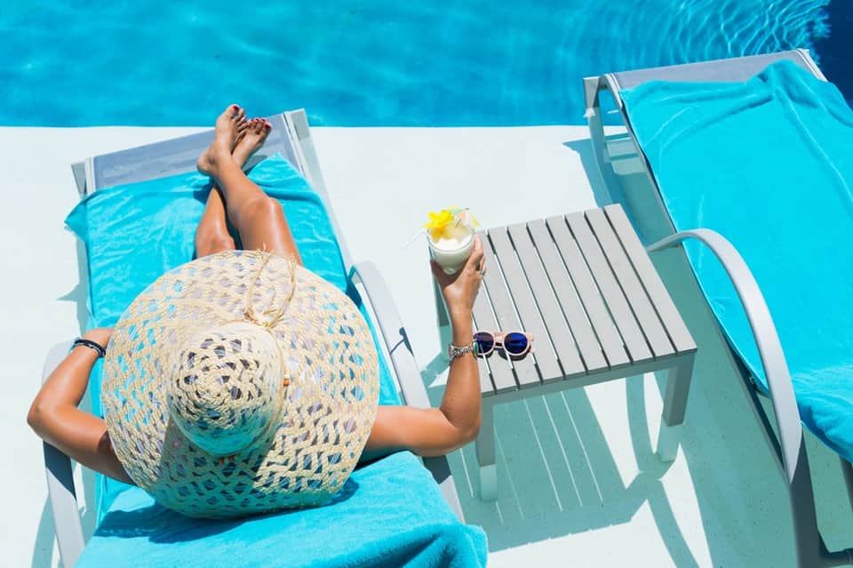 Overhead view of resort guest lounging by bright blue pool with hat and tropical cocktail