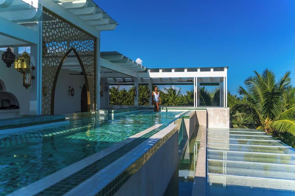 Indoor lap pool with turquoise water, white pergola, and tropical views through glass panels