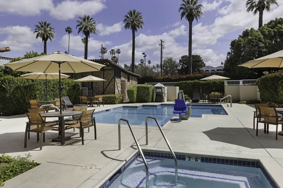 Resort pool area with beige umbrellas, palm trees, spa building, and hot tub in foreground