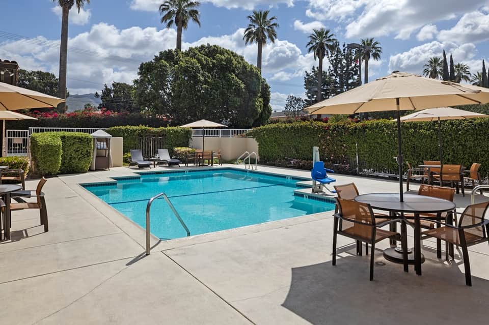 Resort swimming pool with beige umbrellas, lounge chairs, and tropical landscaping with palm trees