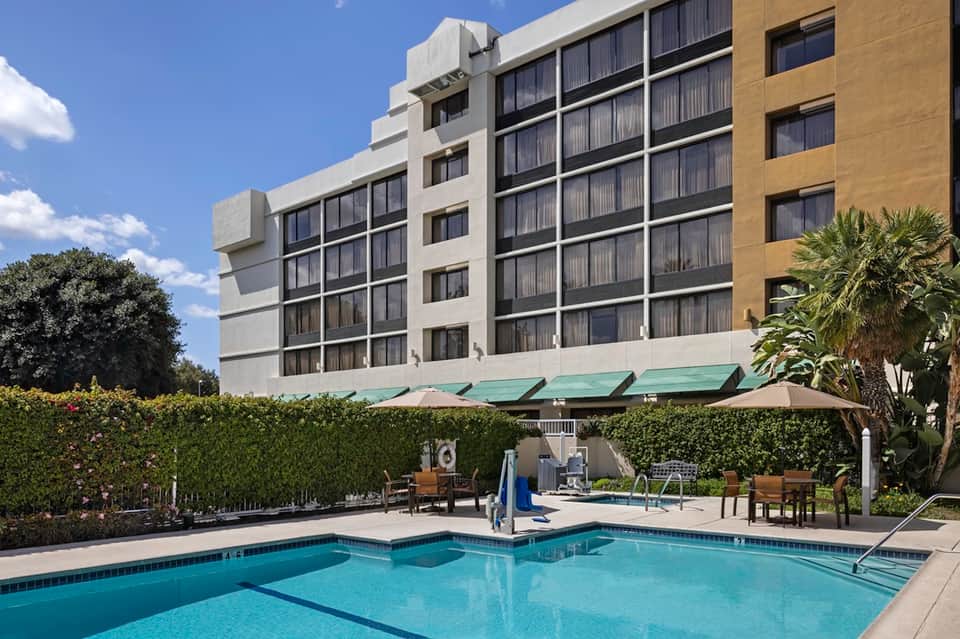 Resort pool area with modern hotel building, hedge border, green umbrellas, and lounge seating