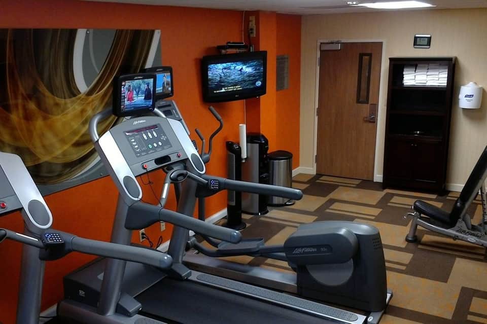 Fitness center with orange walls, treadmill, elliptical machine, wall-mounted TV screens, and wooden accents