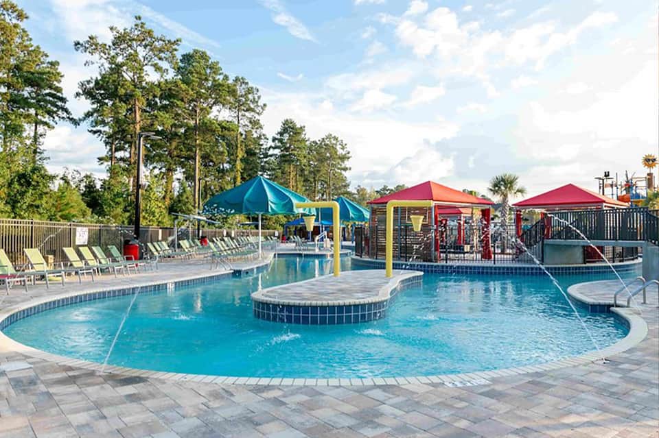 Outdoor resort pool with colorful umbrellas, water features, and lounge chairs surrounded by trees