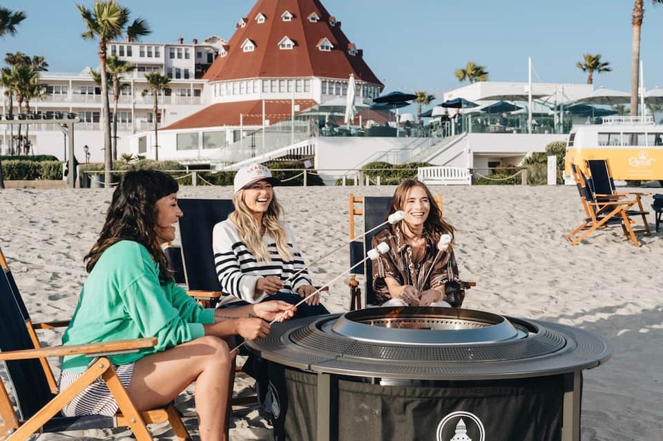 Beachfront guests gathering by fire pit near iconic red-roofed historic building and ocean