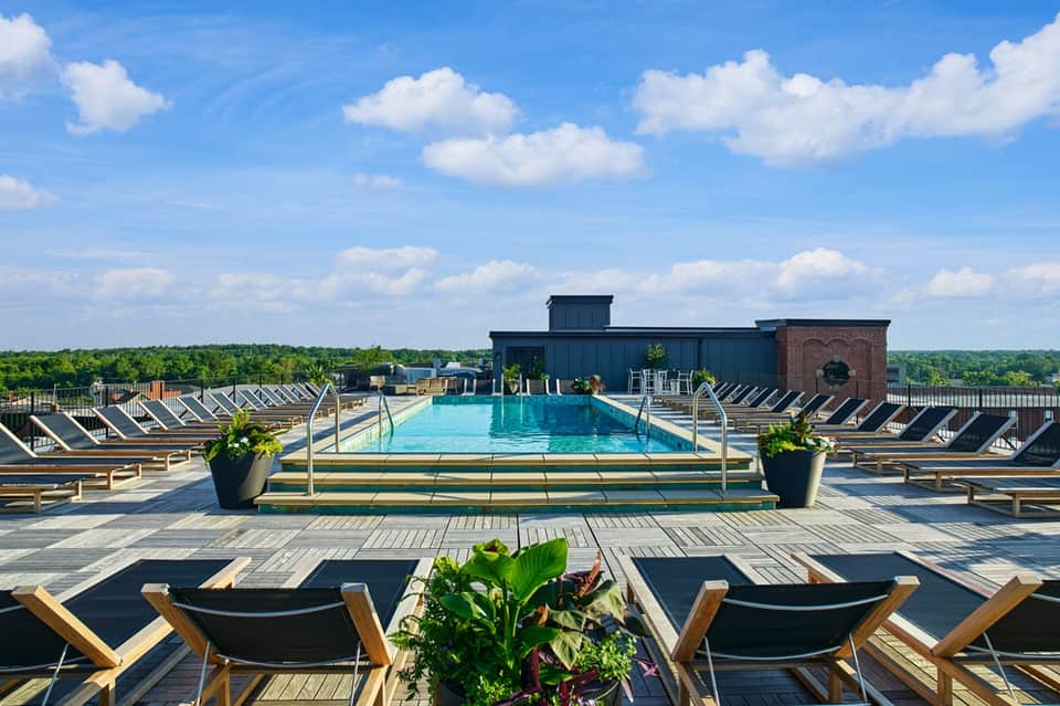 Rooftop pool with blue water, black and wood lounge chairs, cityscape and trees in background