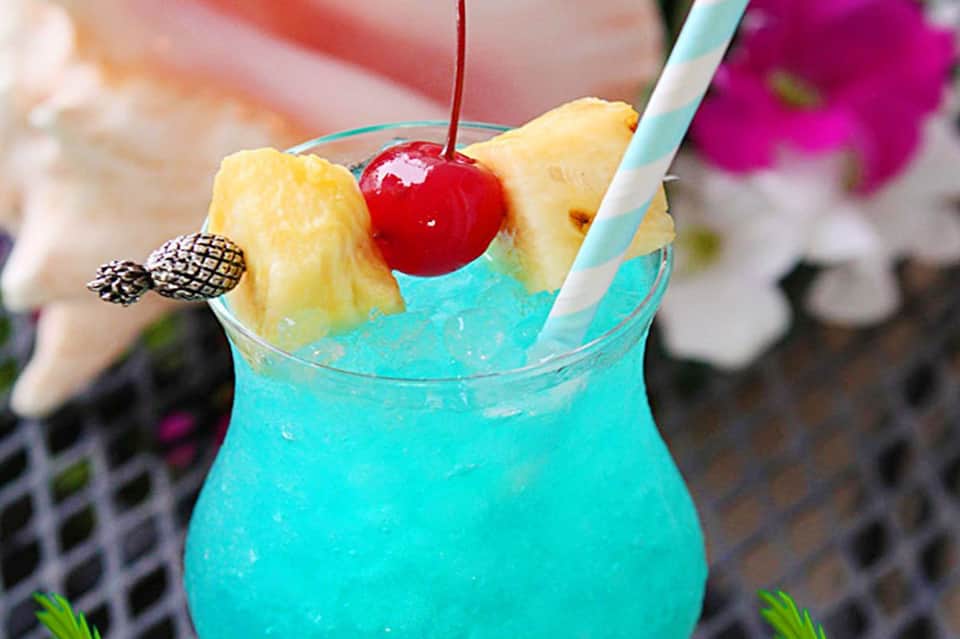 Bright turquoise tropical cocktail garnished with pineapple wedges and a maraschino cherry with striped straw