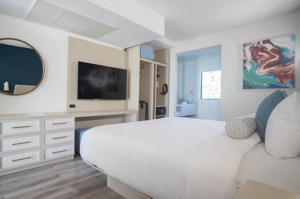 Bright hotel room with white bedding, mounted TV, round mirror, and artwork on walls