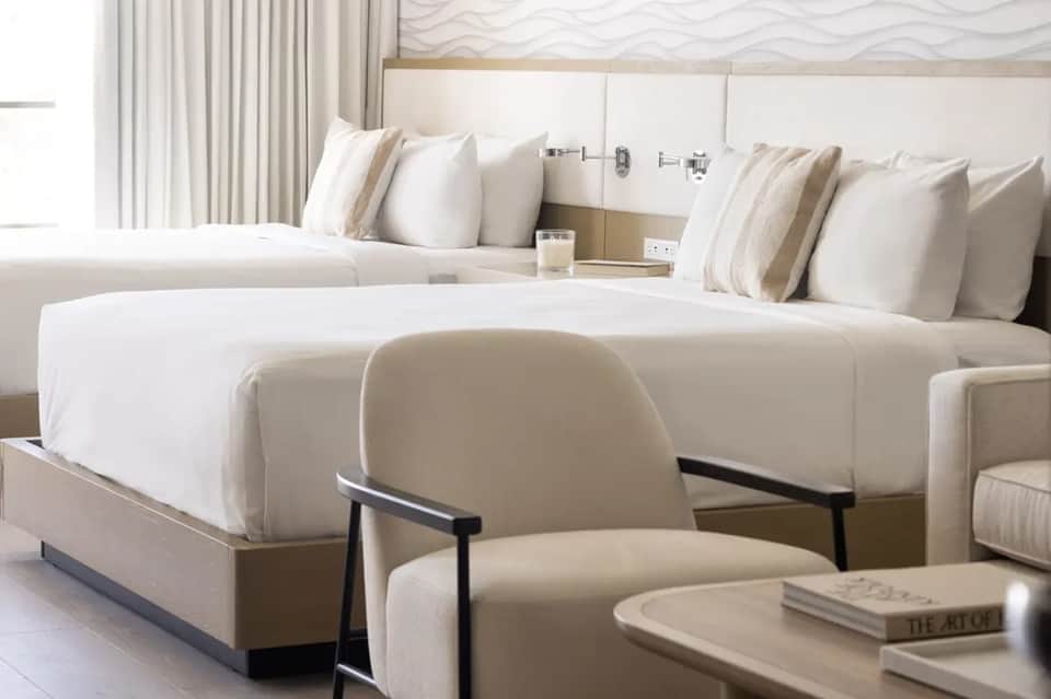 Luxury hotel suite with two queen beds, cream bedding, accent chair, and contemporary decor