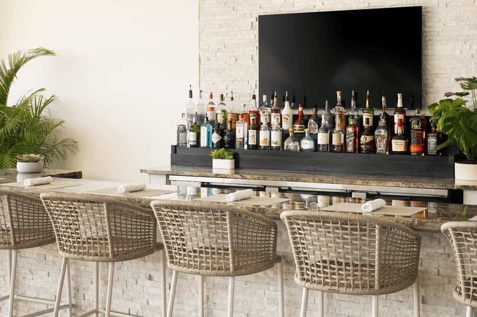 Modern outdoor bar with white wicker stools, extensive liquor selection, and minimalist design