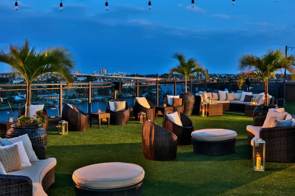 Waterfront lawn lounge at dusk with wicker seating, palm trees, and city bridge view