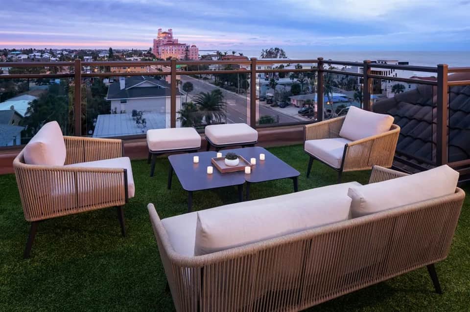 Rooftop lounge with wicker furniture, pink cushions, and city skyline at dusk