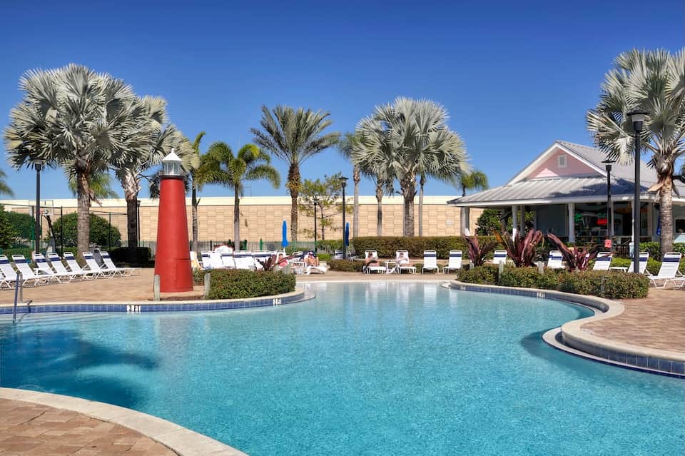 Hotel pool with red lighthouse feature, palm trees, white loungers, and pink clubhouse
