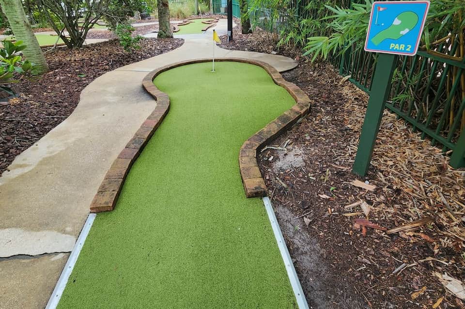Par 2 mini golf hole with bright green synthetic turf and brick border in tropical setting