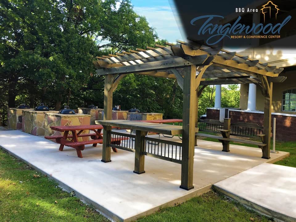 BBQ area at resort with wooden pergola, picnic tables, and grills surrounded by trees