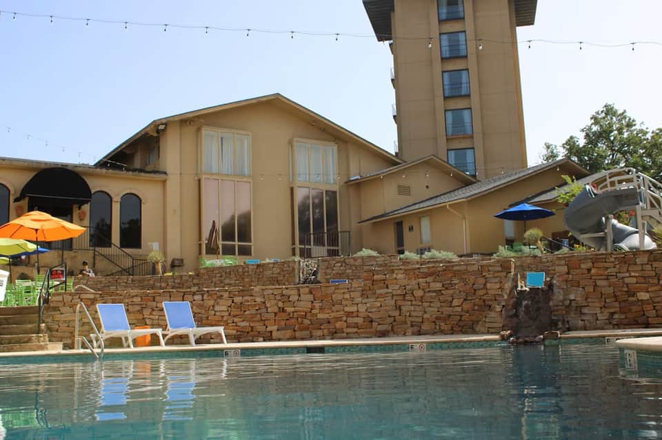 Resort swimming pool with stone retaining wall, lounge chairs, umbrellas, and multi-story hotel tower