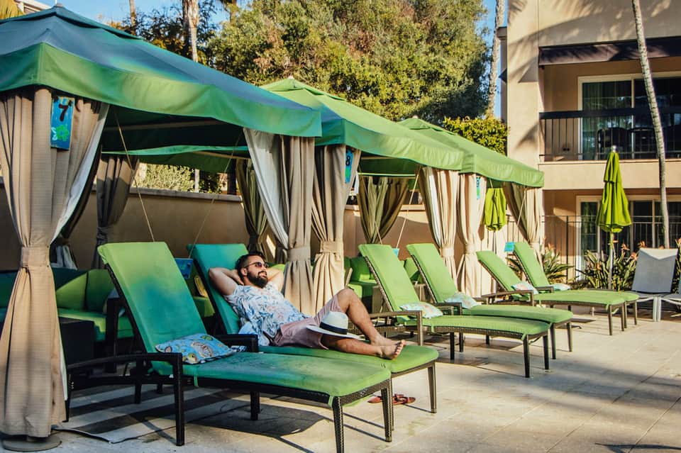 Man relaxing on green lounge chair under cabana with beige curtains at resort pool deck