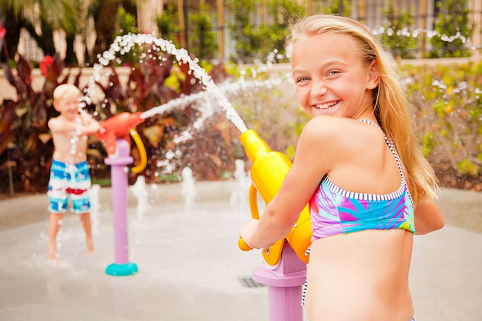 Children playing in a splash pad with water fountains and tropical plants in background