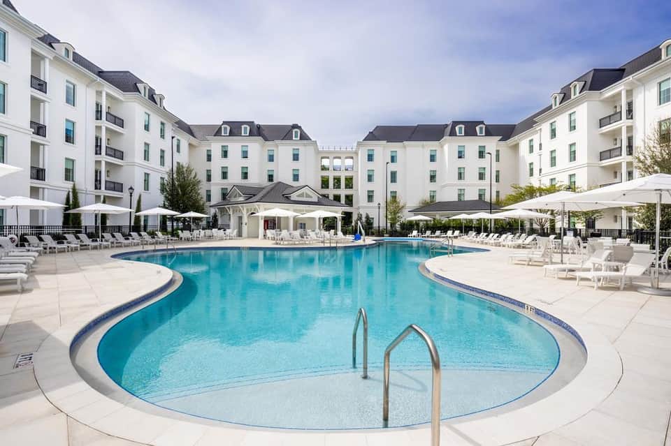 Resort pool courtyard with blue water, white lounge chairs, umbrellas, and elegant white residential buildings