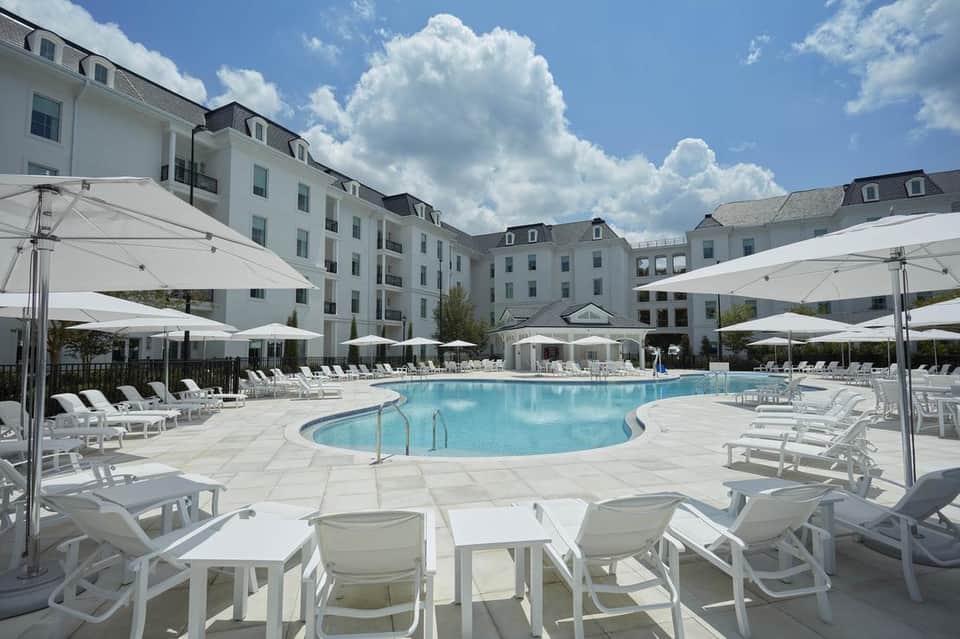 Curved resort pool surrounded by white loungers, umbrellas, stone patio, and colonial-style hotel building