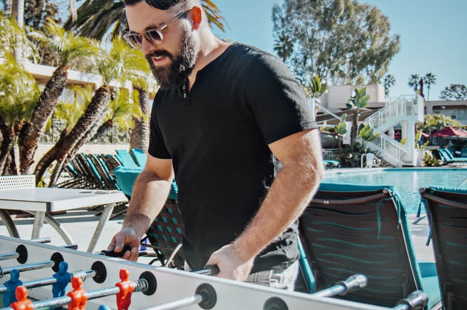 Man playing foosball by resort pool with palm trees and lounge chairs on sunny day