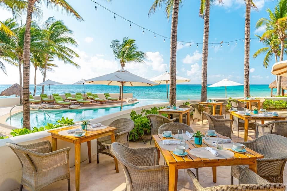 Beachfront dining area with woven chairs, wooden tables, palm trees, ocean view, and string lighting
